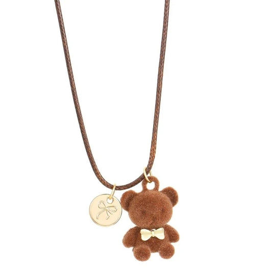 A brown cord necklace featuring a small brown plush teddy bear charm with a gold bow tie and a round gold disc charm engraved with a bow, shown against a plain white background.