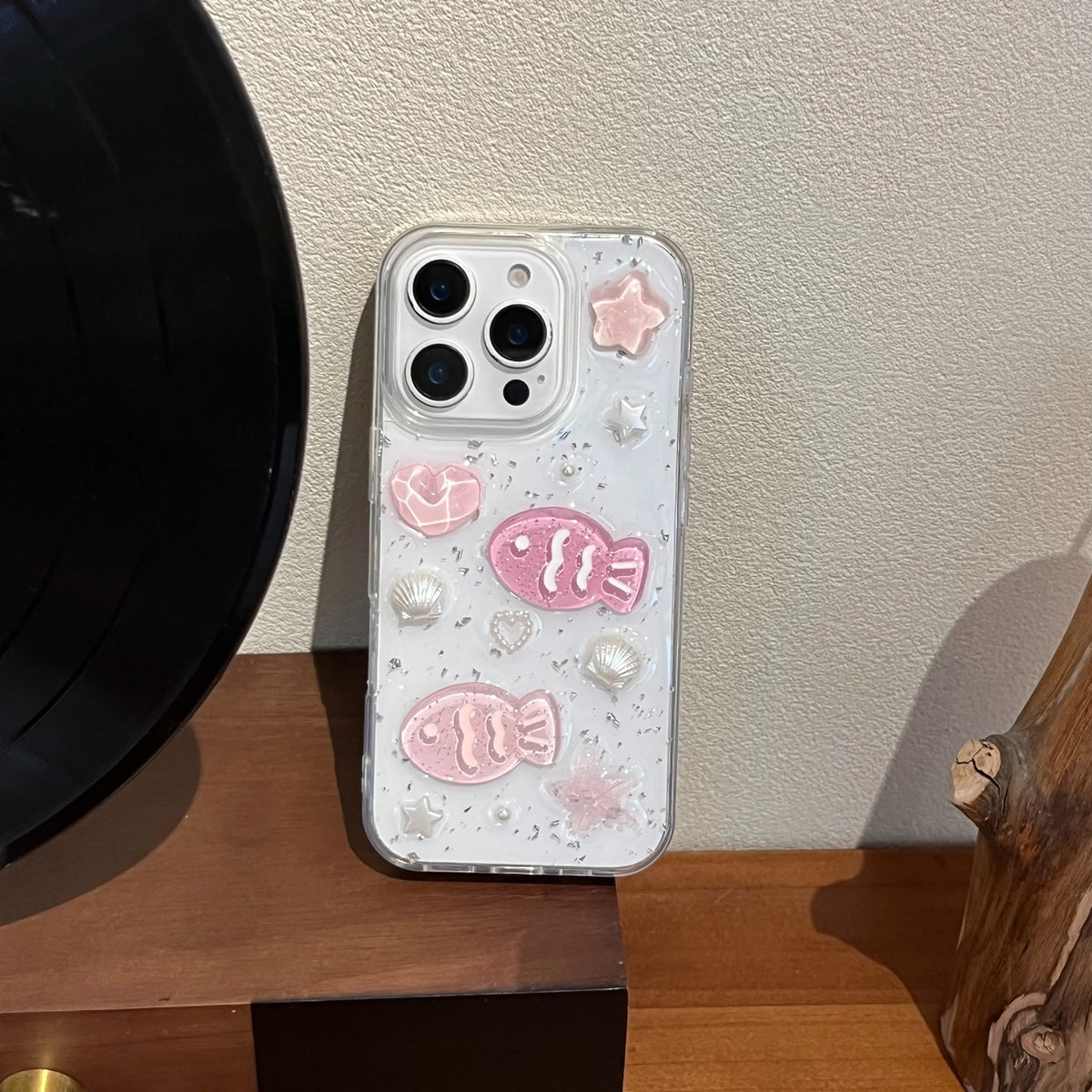 A clear phone case decorated with pink 3D fish, pearls, and glitter, shown on a phone resting on a wooden surface next to a black vinyl record.