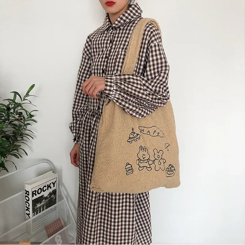 A person wearing a brown and beige gingham shirt and matching pants carries a beige sherpa tote bag with black cartoon bunny drawings and the word 'MATE' on a plain white wall.