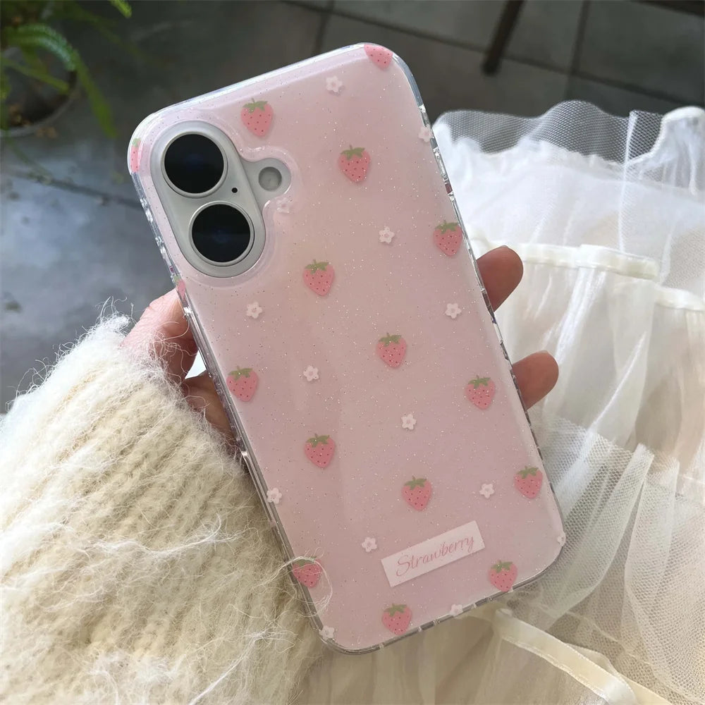 A hand wearing a cream-colored fuzzy sweater holds a clear phone case decorated with pink strawberries and small white flowers on a light pink background. The case has a small white label near the bottom that reads 'Strawberry'.
