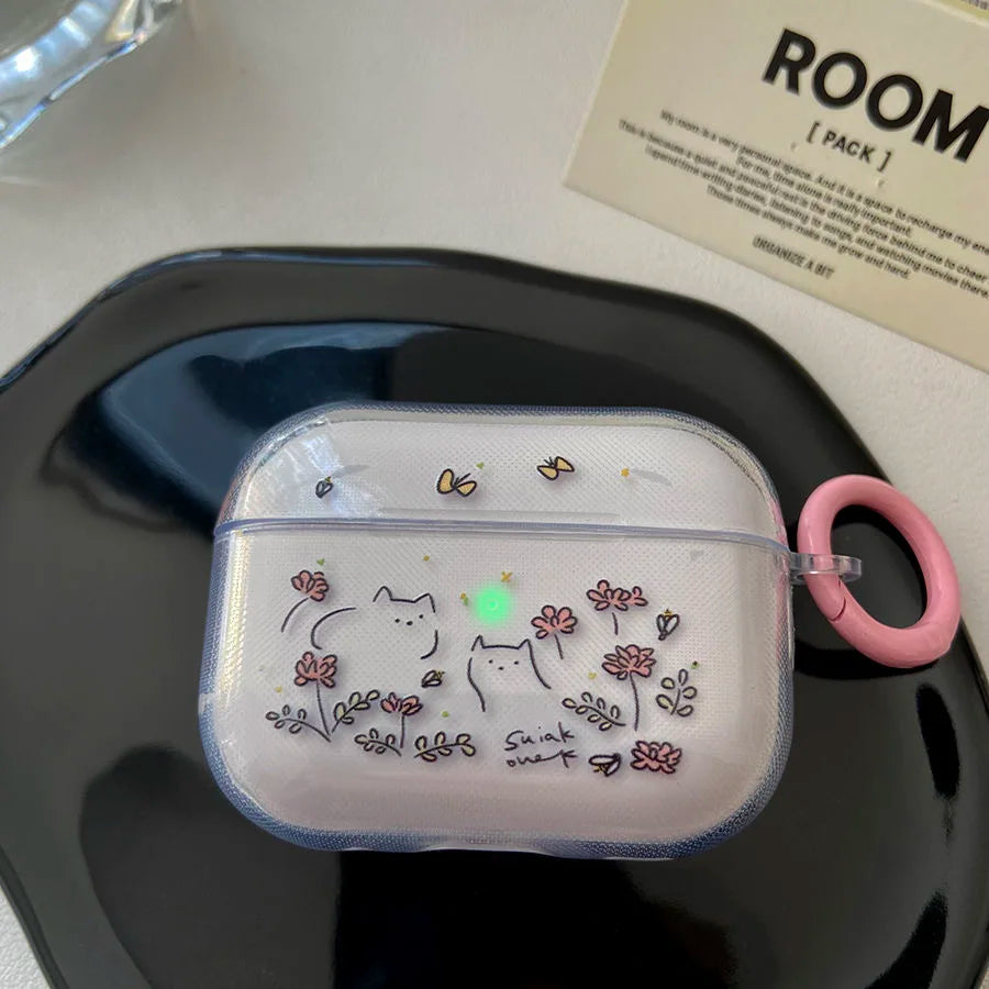 A clear protective case for white AirPods, decorated with cute cartoon cat illustrations, small pink flowers, and a pink carrying loop, resting on a black tray.