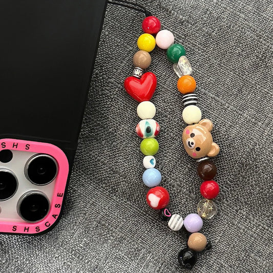 A colorful beaded phone strap with a red heart, a bear charm, and patterned beads, shown next to a black phone case with a pink camera border on a gray textured fabric background.