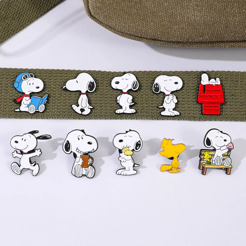 Ten Snoopy enamel lapel pins displayed on a white surface, with a matching olive green canvas strap laid above them. The pins feature Snoopy in various poses and scenes, including reading a book, sitting, standing, with Woodstock, and in a red doghouse scene.