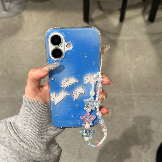A hand holding a blue glittery phone case decorated with white bunny illustrations and a pastel beaded charm strap with star and bunny details. The case has a clear bumper edge and is shown against a gray concrete floor background.