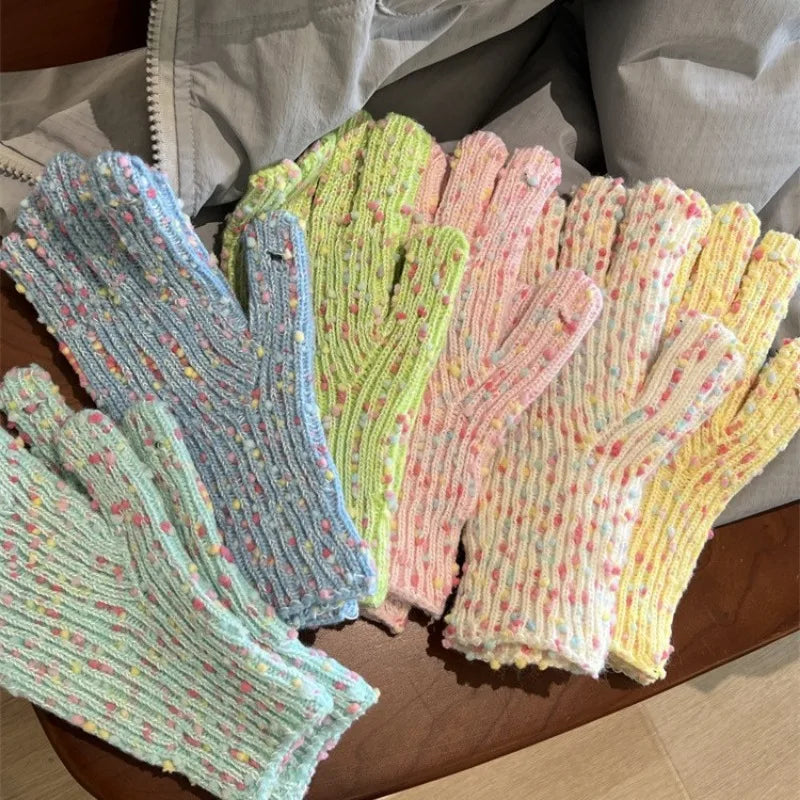 Four pairs of chunky knit gloves in pastel colors with tiny speckled flecks, laid out on a wooden surface with a gray jacket in the background.