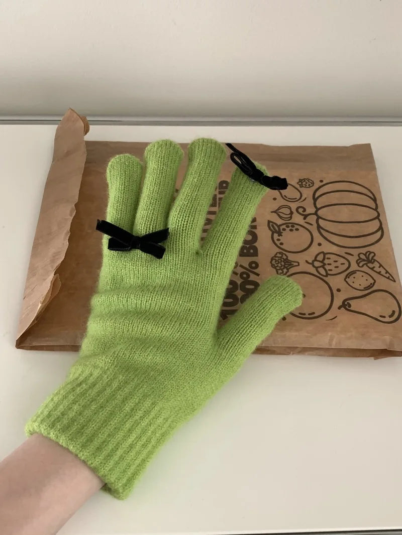 Green knitted glove with a black bow on a white surface with a brown paper bag in the background.