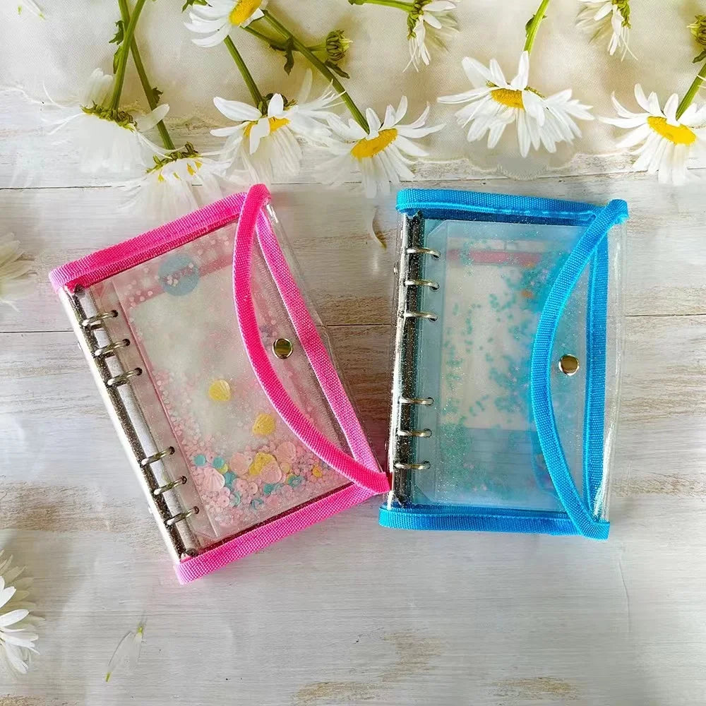 Two small clear binder sleeves with ribbon trim, one pink and one blue, shown on a light wood surface with white daisies around them.