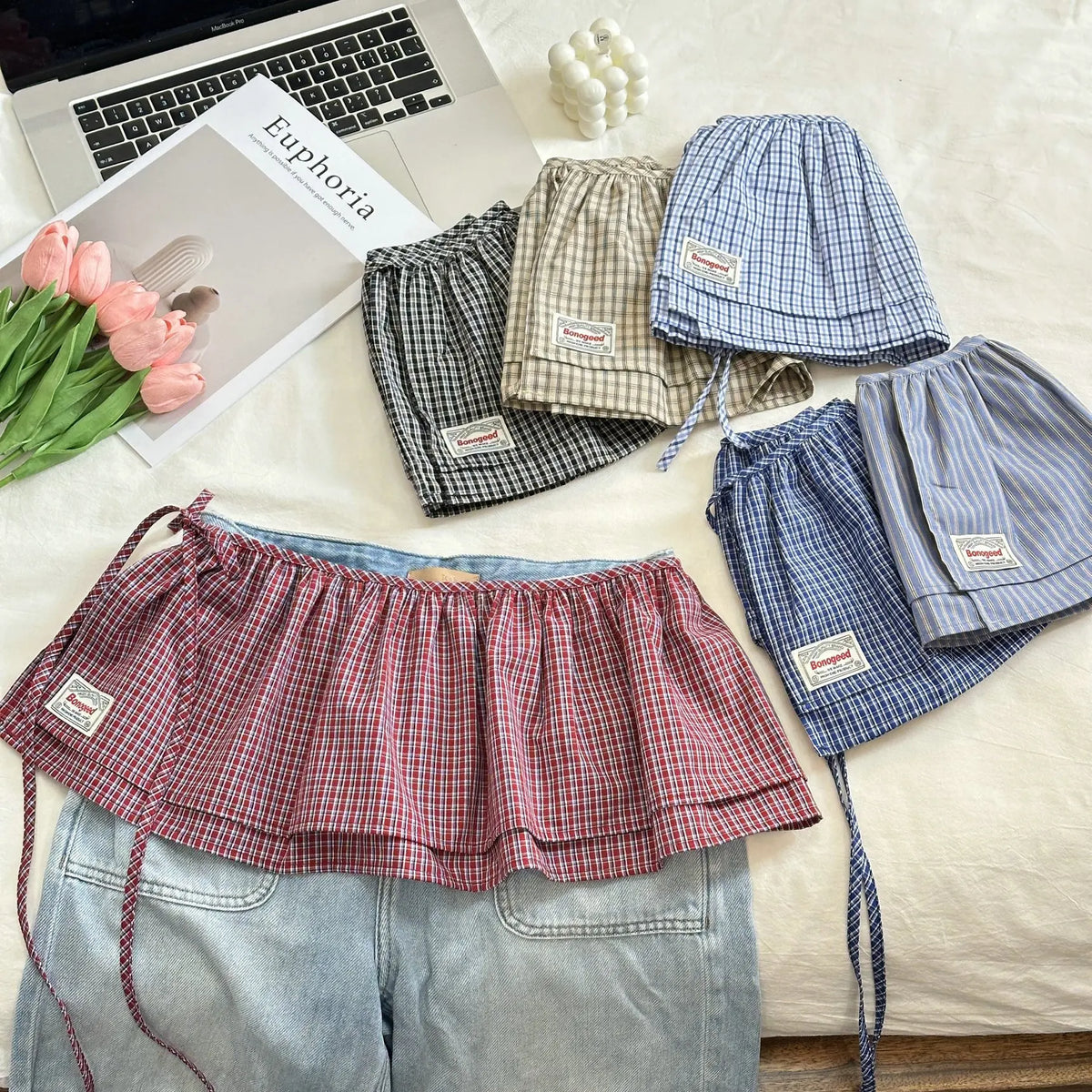 Plaid skirts and shorts displayed on a bed with a laptop and flowers.