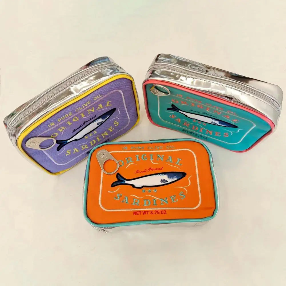 Three rectangular zippered pouches designed to look like sardine tins, arranged on a plain light background. One tin has a purple lid with yellow trim, one has a teal lid with pink trim, and one has an orange lid with teal trim.