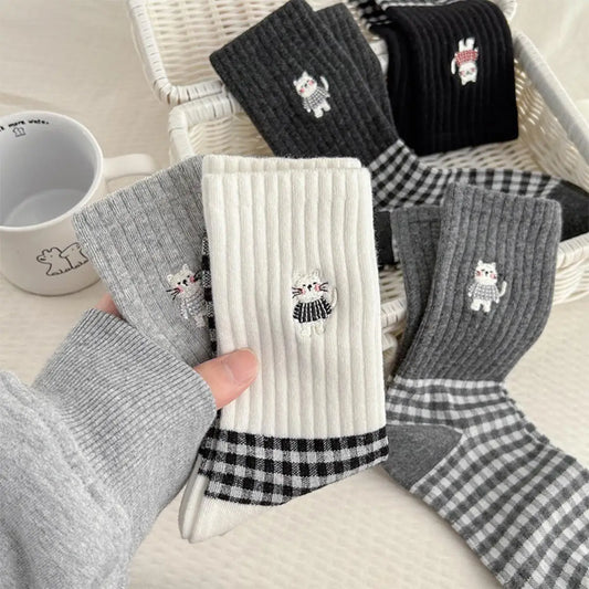 A hand holds a white ribbed sock with a black-and-white cat patch and black gingham cuff, while several other socks in gray, black, and white gingham are arranged in a white basket beside a white mug.