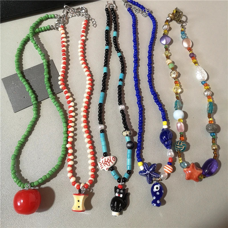 Six colorful beaded necklaces with unique charms are displayed side-by-side on a neutral gray surface. The necklaces feature green, red-and-white, black-and-turquoise, royal blue, and multicolor bead strands with charms including a red apple, a small house, a black cat, a blue owl, a starfish, and a blue egg.
