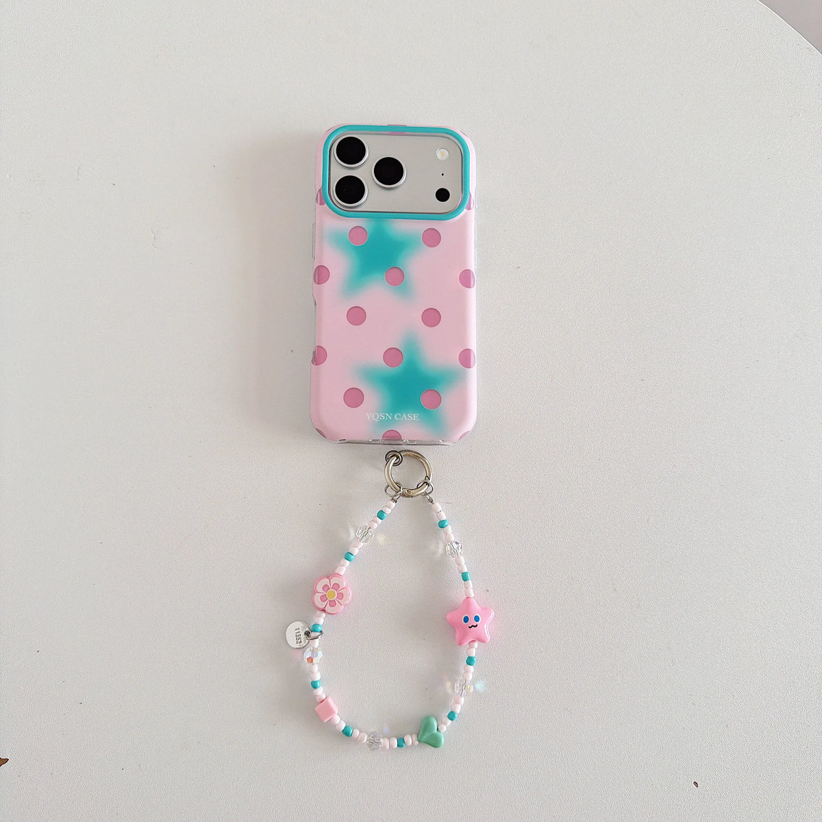 A pink phone case with turquoise polka dots and star patterns, attached to a matching beaded phone strap with a small charm, displayed on a plain white surface.