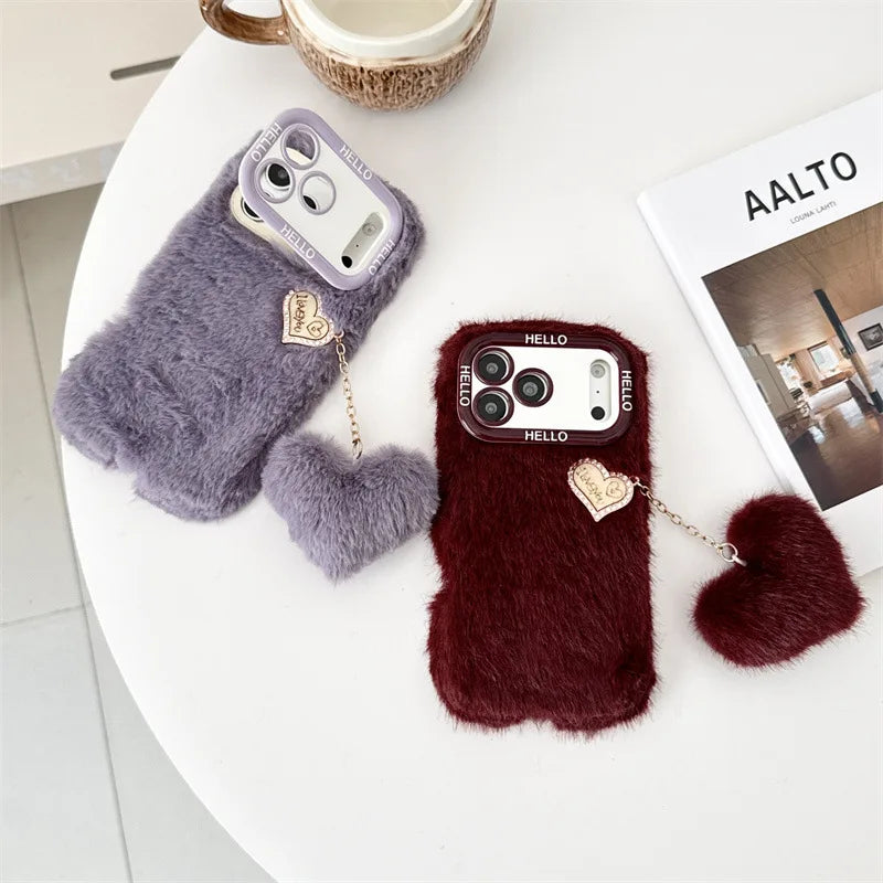 Two fuzzy faux-fur phone cases on a white table, one lavender and one deep burgundy, each with a small heart charm and chain.