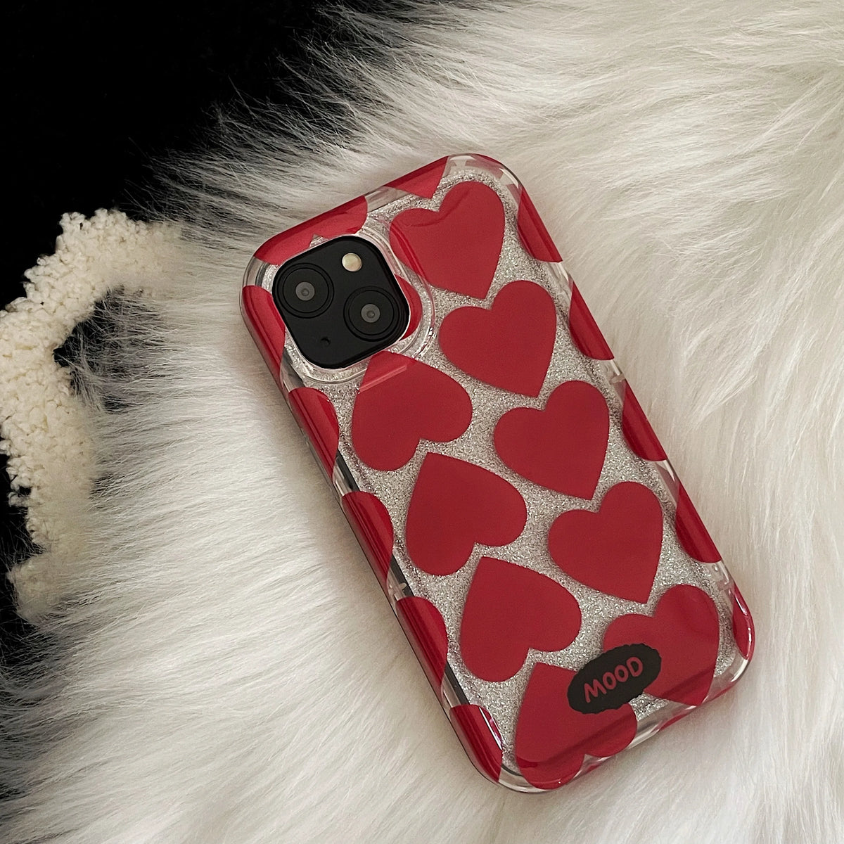 A clear phone case with a pattern of red hearts over a silver glitter background, shown on an iPhone. The case has a black bumper edge and a small black oval logo at the bottom that reads 'MOOD'.