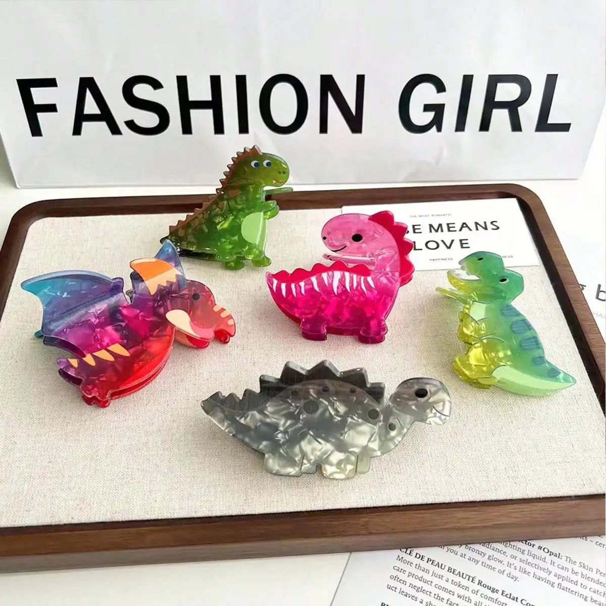 Colorful dinosaur-shaped hair clips on a tray with 'Fashion Girl' text in the background.