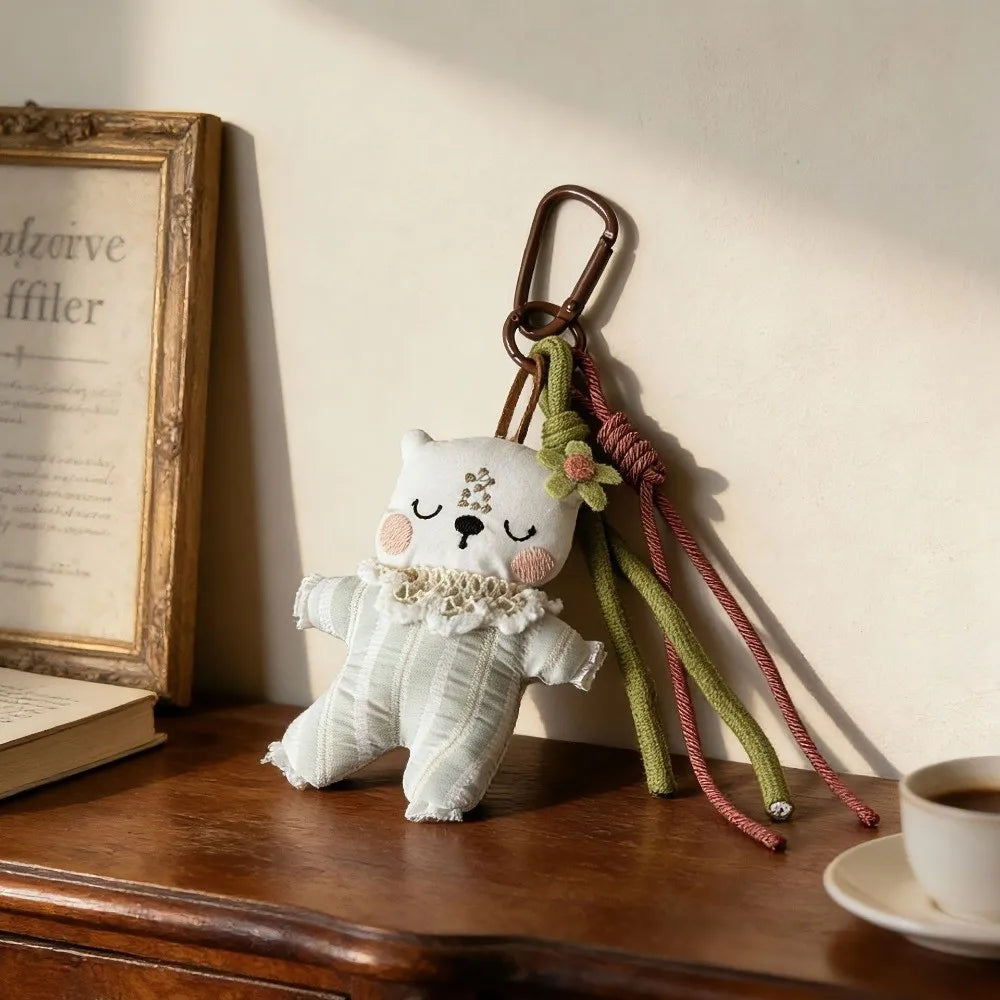 A small plush bear keychain or bag charm in soft off-white fabric with embroidered facial details and a ruffled trim, hanging from a metal carabiner on a wooden table. The scene includes a framed print, an open book, and a cup of coffee in a warm, styled interior setting.