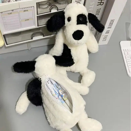 Two plush dog-shaped pencil cases on a gray desk, one standing and one lying down with a zippered pocket showing pens inside. The dogs are white with black patches.