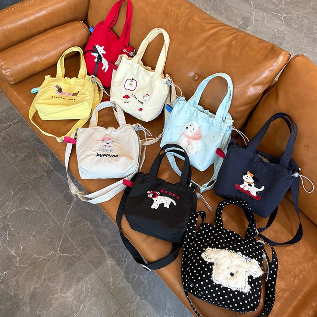 Seven small tote bags in different colors are arranged on a brown leather couch. The bags feature cartoon dog illustrations and embroidered text, with some having white faux-fur dog patches on the front.
