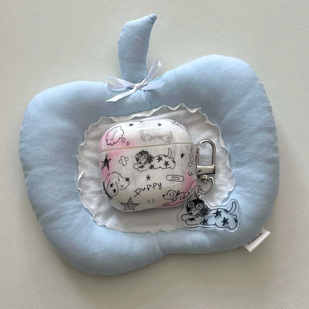 A light blue plush cushion shaped like an apple, styled as a decorative prop. In the center sits a white fabric AirPods case decorated with black doodles of dogs, stars, and the word 'Puppy', complete with a silver metal clasp and a small matching dog charm.