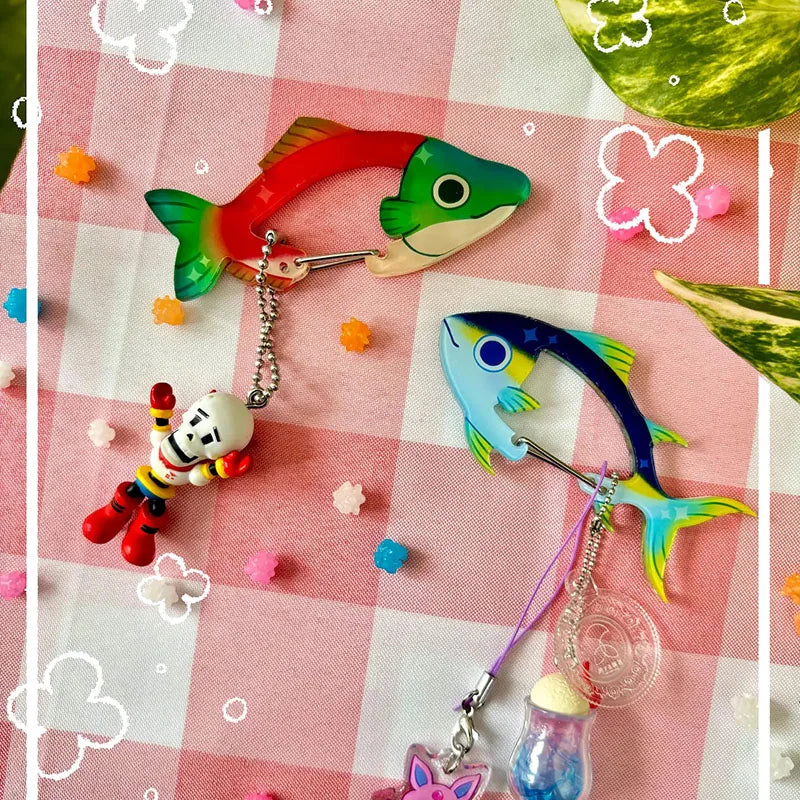 Two colorful fish-shaped carabiner clips with dangling charms, displayed on a pink and white checkered blanket with small decorative flower stickers.