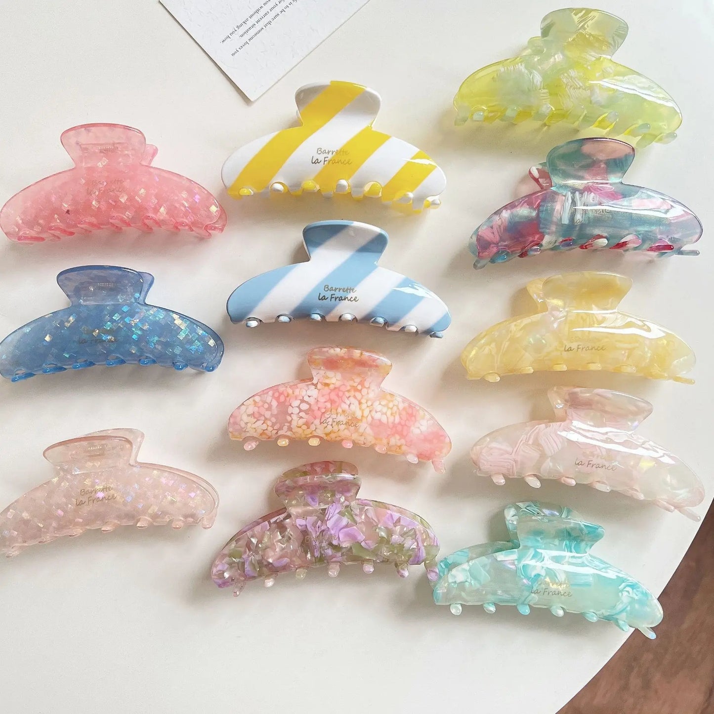 Twelve translucent claw hair clips in assorted pastel colors and patterns, including floral prints, stripes, glitter, and marble textures, arranged on a white surface.