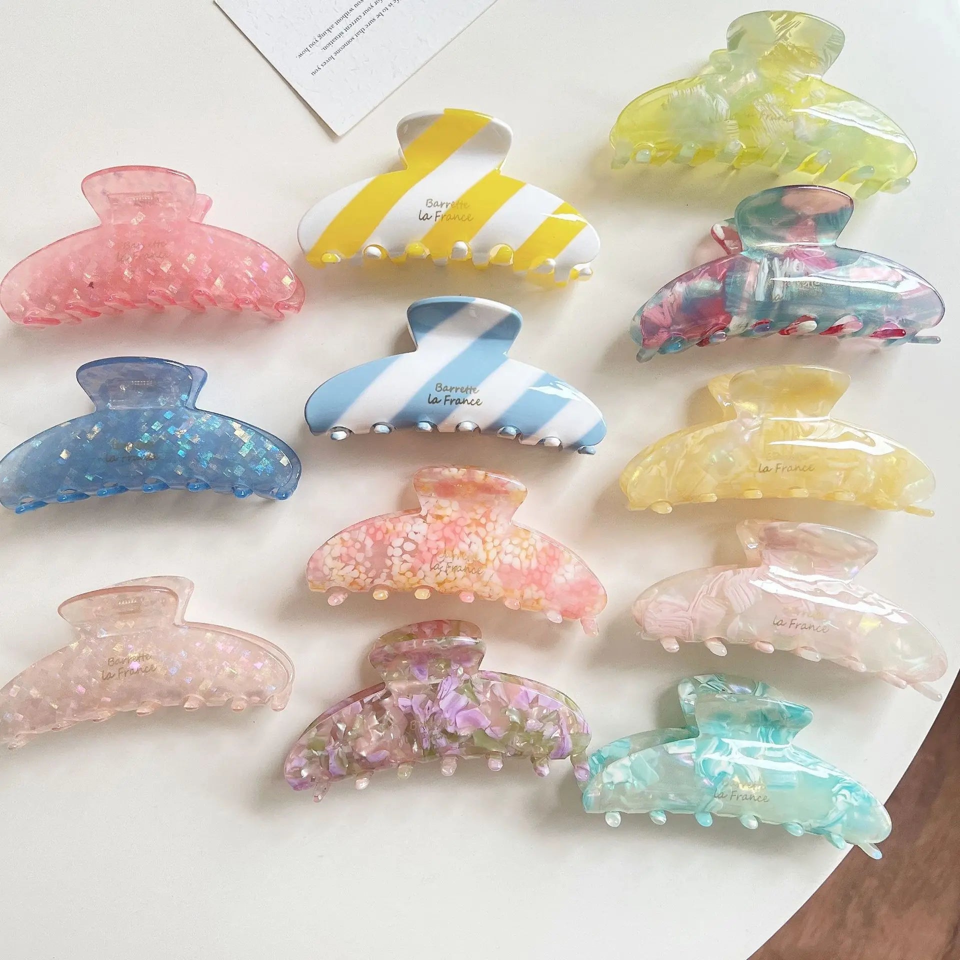 Twelve translucent claw hair clips in assorted pastel colors and patterns, including floral prints, stripes, glitter, and marble textures, arranged on a white surface.