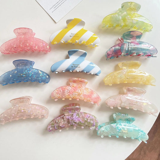 Twelve translucent claw hair clips in assorted pastel colors and patterns, including floral prints, stripes, glitter, and marble textures, arranged on a white surface.