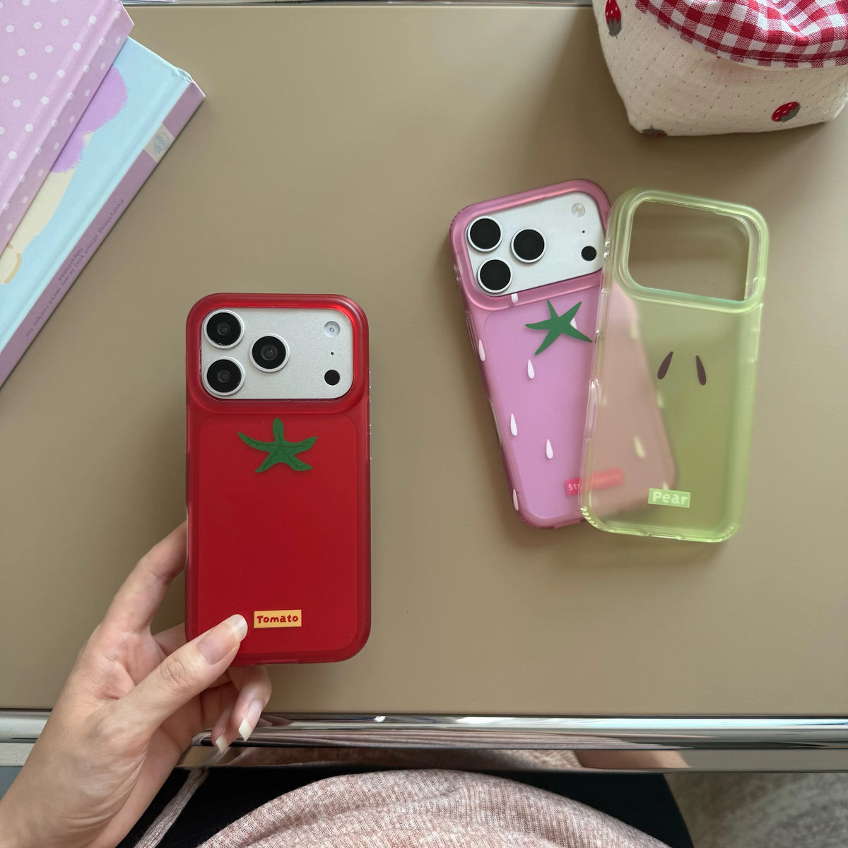 Three phone cases with cute cartoon designs are displayed on a beige tabletop. One red case is held by a hand, while two more cases in pink and translucent light green rest on the table.
