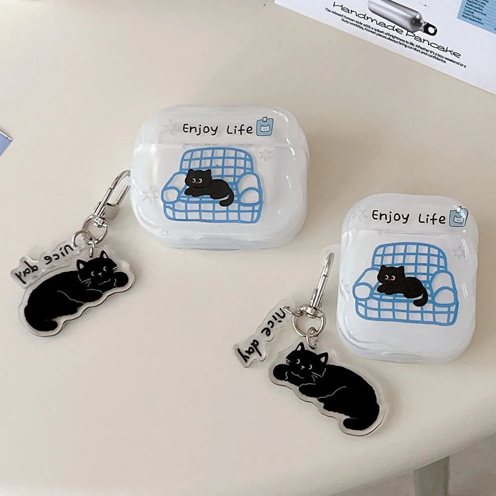 Two clear AirPods case covers with a blue couch graphic and black cat illustration, each attached to a black cat keychain charm with silver hardware. The cases are placed on a light beige surface with a partial paper card visible in the top right.