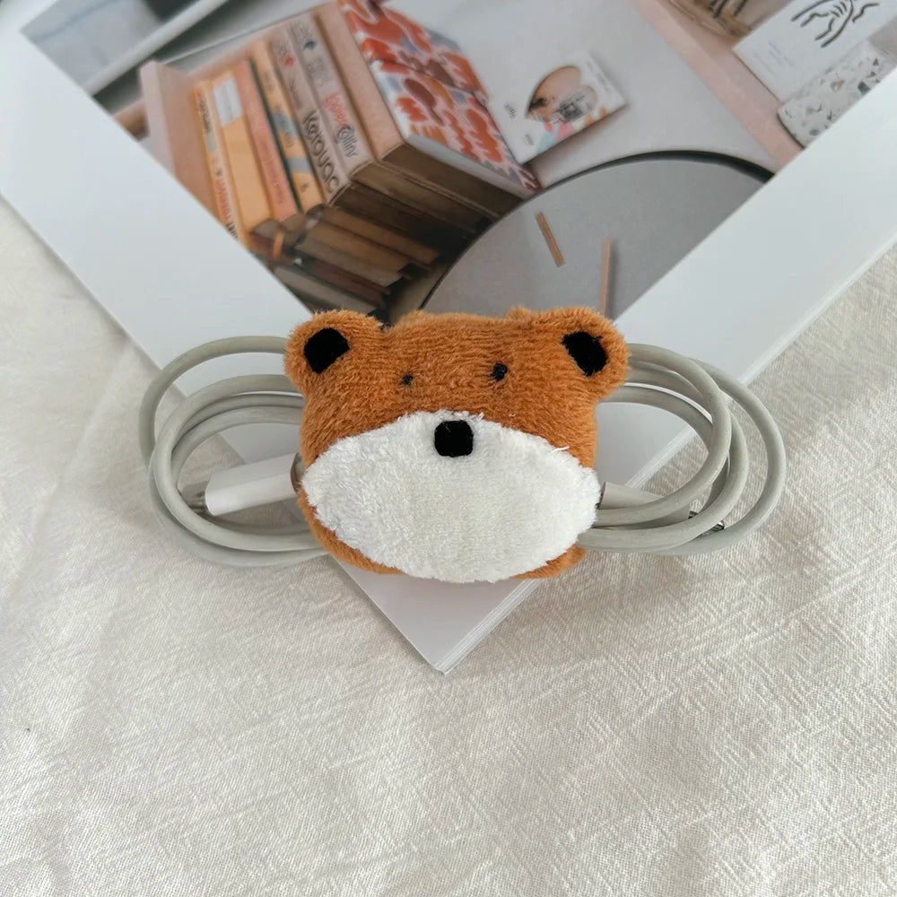 A plush fox-head cable tie or cable organizer resting on a white textured fabric surface, with coiled gray cables wrapped around it and a framed picture in the background.
