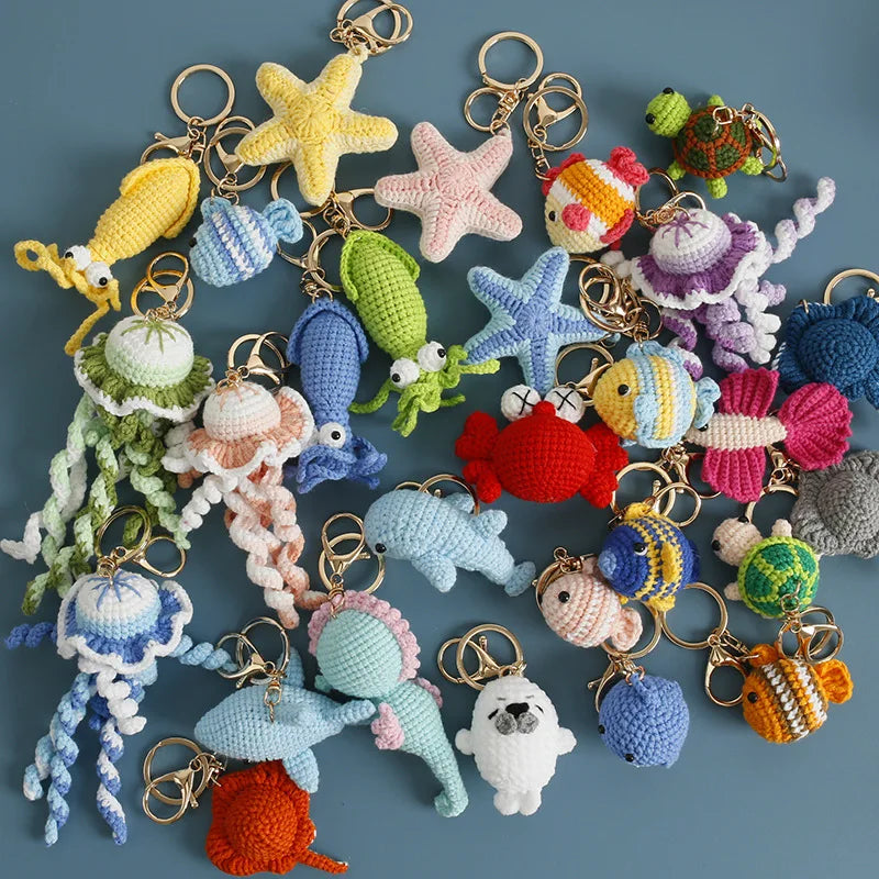 A collection of colorful crocheted sea creature keychains, including starfish, octopus, jellyfish, crab, fish, and turtle designs, arranged on a solid blue-gray background.