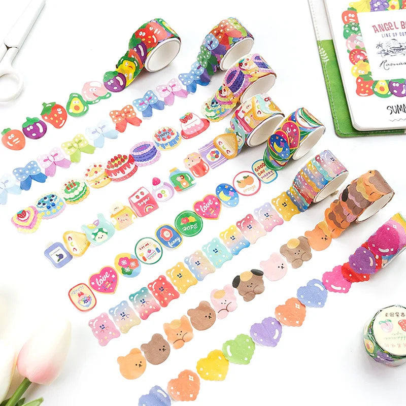 A flat lay of multiple rolls of decorative washi tape with colorful cartoon and food illustrations, arranged on a white surface with a notebook, scissors, and a tulip as props.