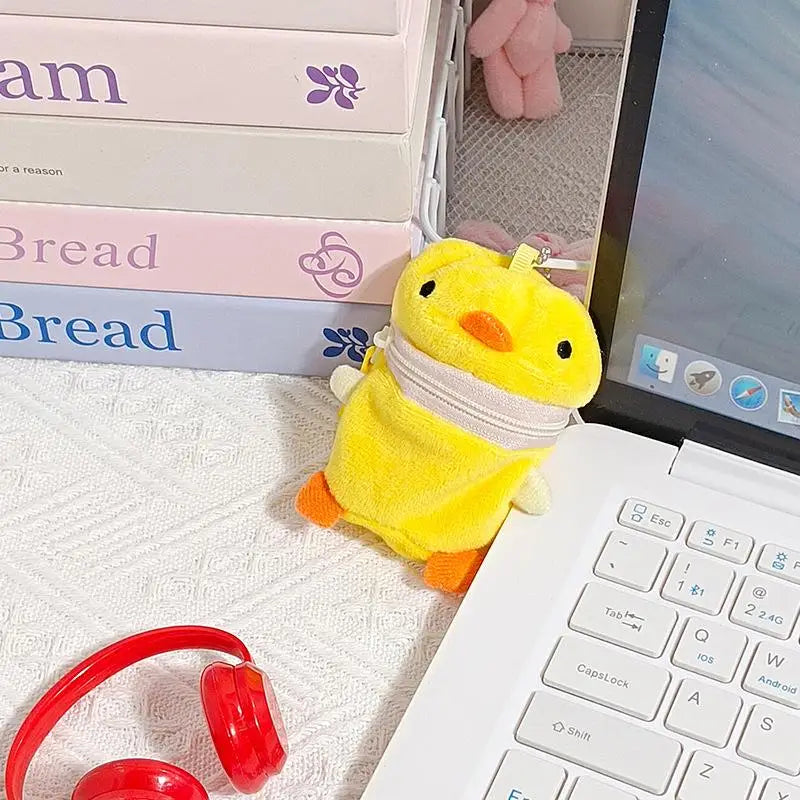 A yellow plush duck-shaped pouch with an orange beak and zipper closure, placed on a white textured surface next to a white laptop, a stack of pastel books, and red headphones.