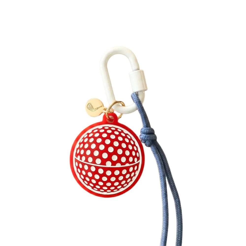 A red and white polka dot golf ball keychain or bag charm with a white carabiner, a blue cord, and a small gold tag on a plain white background.