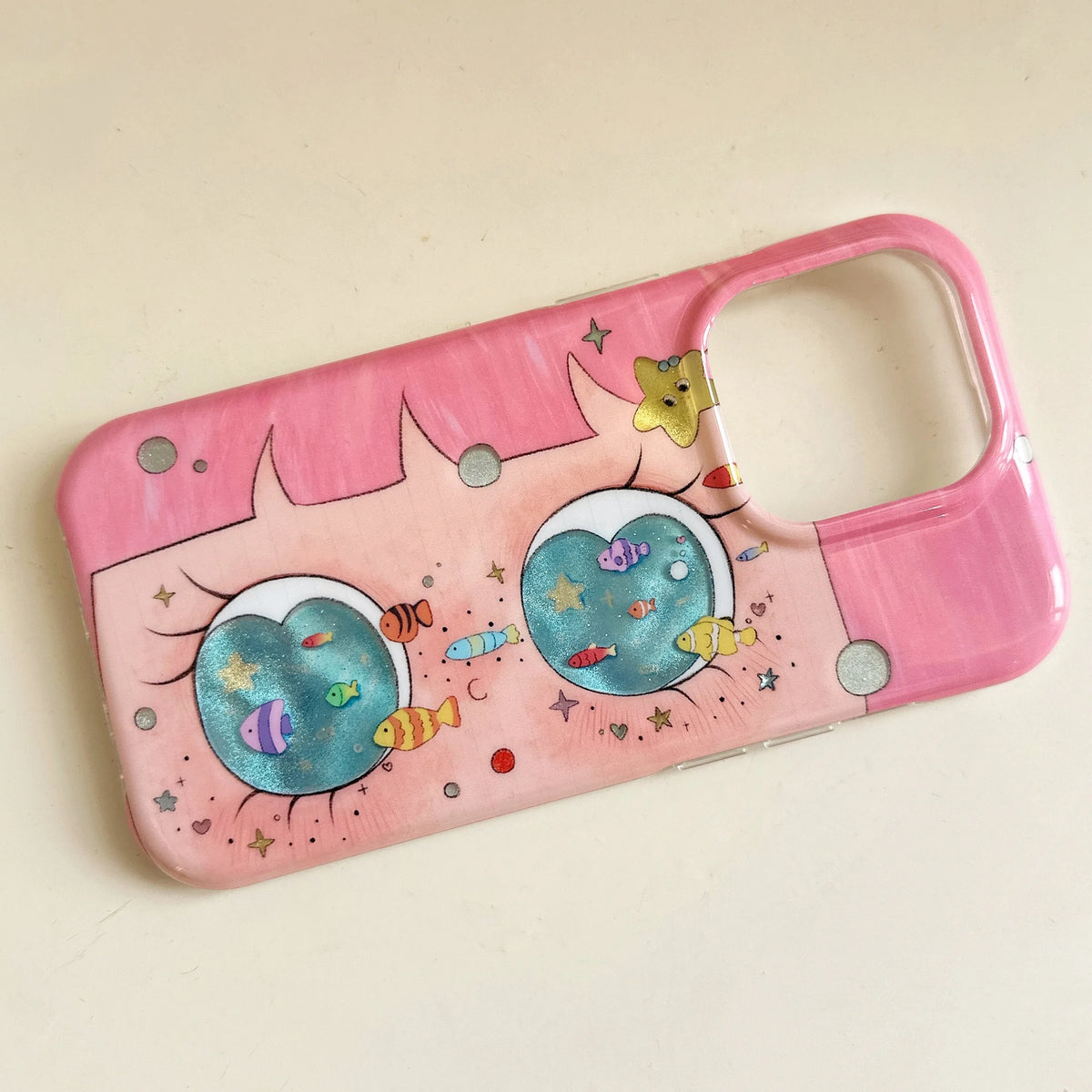 A pink phone case with a whimsical cartoon design featuring two large eyes, colorful fish, and stars, photographed on a light beige background.