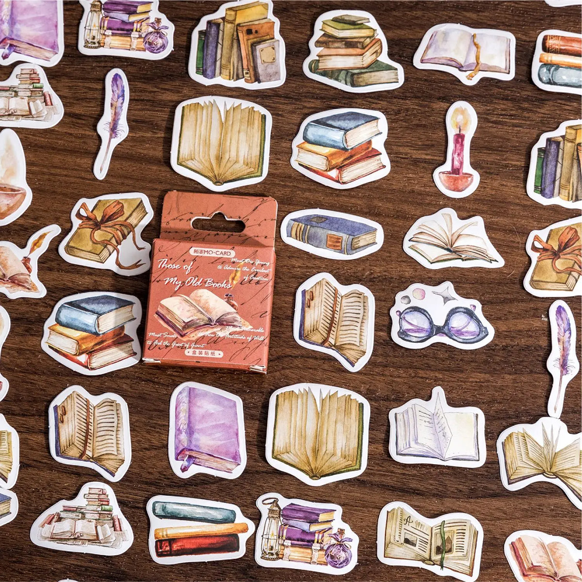 A sheet of die-cut stickers featuring watercolor illustrations of books, candles, glasses, and quills, displayed on a dark wood background with a small orange package of the stickers in the center.