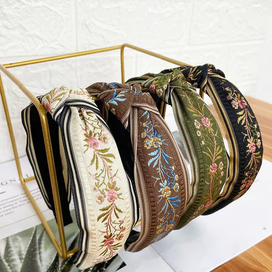 Five wide fabric headbands with floral embroidery and striped trim, displayed on a gold metal stand. The headbands are in cream, black, brown, olive green, and black, each featuring colorful floral patterns and striped accents.