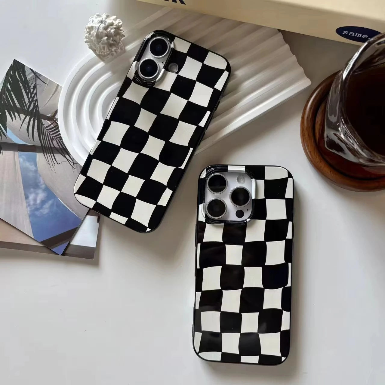 Two black and white checkered phone cases displayed on a white surface with styled props, including a coffee cup, a small decorative object, and a magazine.