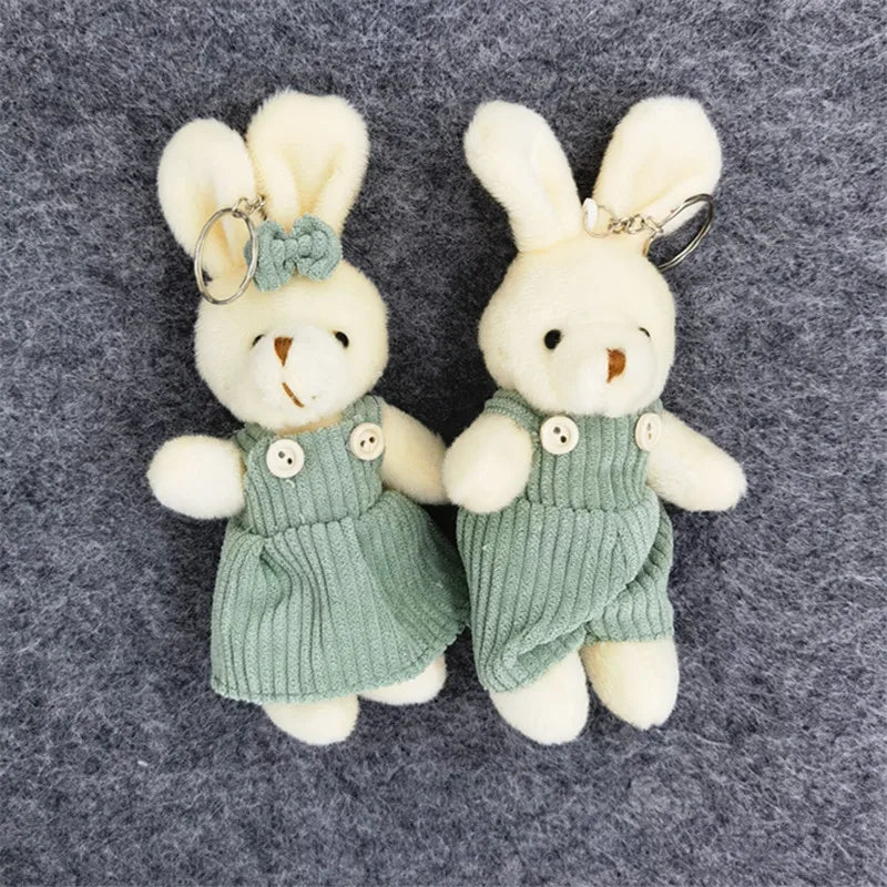 Two small plush bunny keychains dressed in light green corduroy-style outfits, placed side by side on a dark gray textured background.