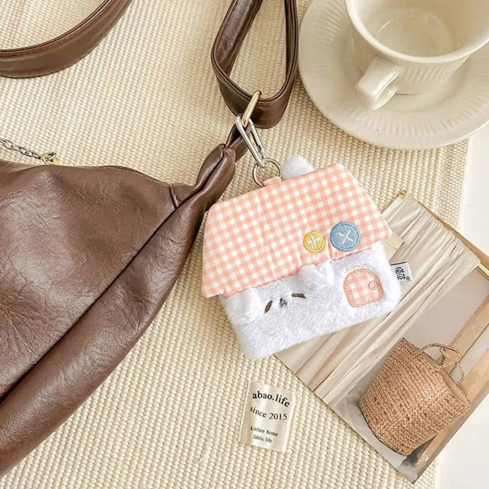 A cute house-shaped plush keychain bag charm with a white fuzzy body, pink-and-white gingham roof, and embroidered button details, shown hanging from a brown handbag strap on a woven beige surface with a cup and book nearby.