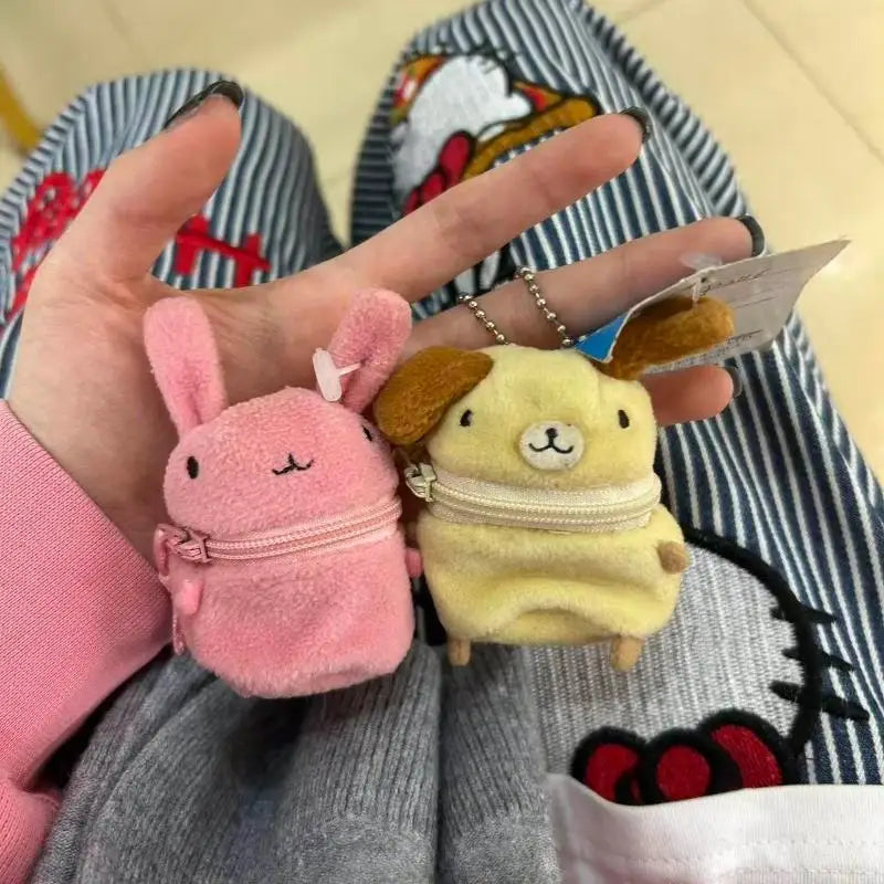 Two small plush character zip pouches held in a hand. One is pink with a bunny face and bow, and the other is pale yellow with a dog face and brown ears.