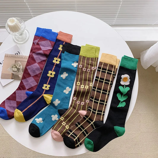 Six pairs of decorative knee-high socks arranged on a round white table. The socks feature various patterns including argyle, floral, plaid, and solid colors in blue, brown, black, teal, purple, and green, with yellow, pink, and white accents.