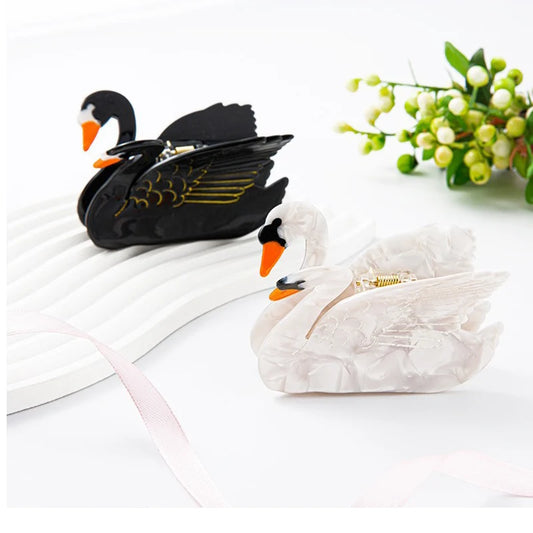Two swan-shaped hair clips, one black and one white, on a white surface with a small plant in the background.