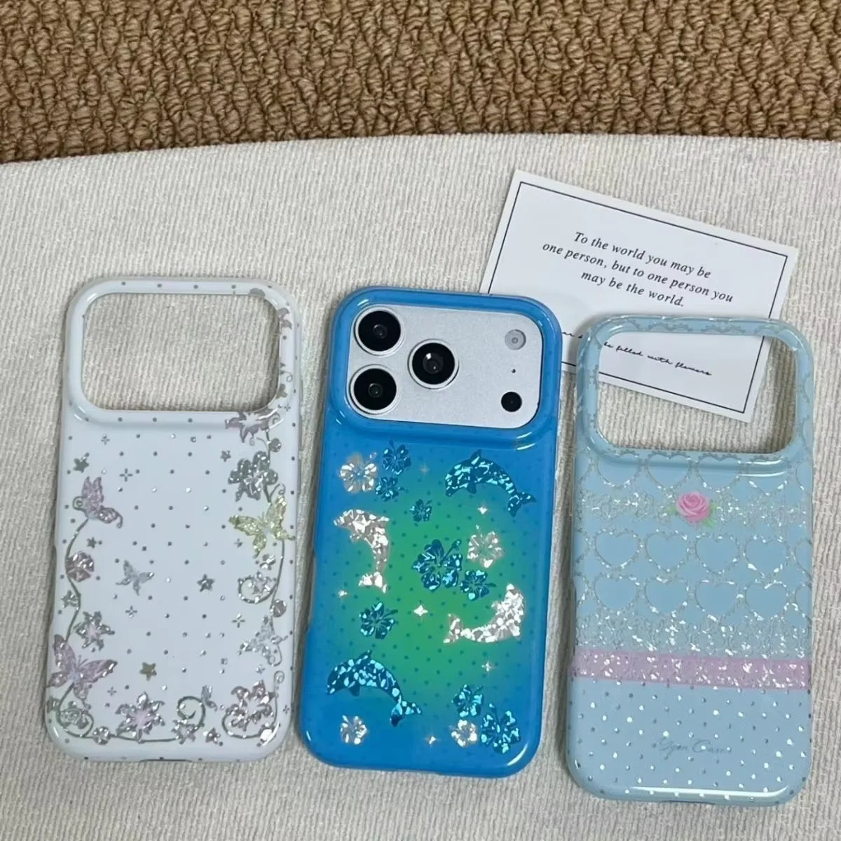 Three decorative smartphone cases arranged on a light fabric surface with a woven brown mat behind them. The left case is white with pastel butterfly and star glitter patterns; the center case is blue with iridescent glitter and a green-to-blue gradient; the right case is pale blue with a pink glitter band and small pink butterfly accents.