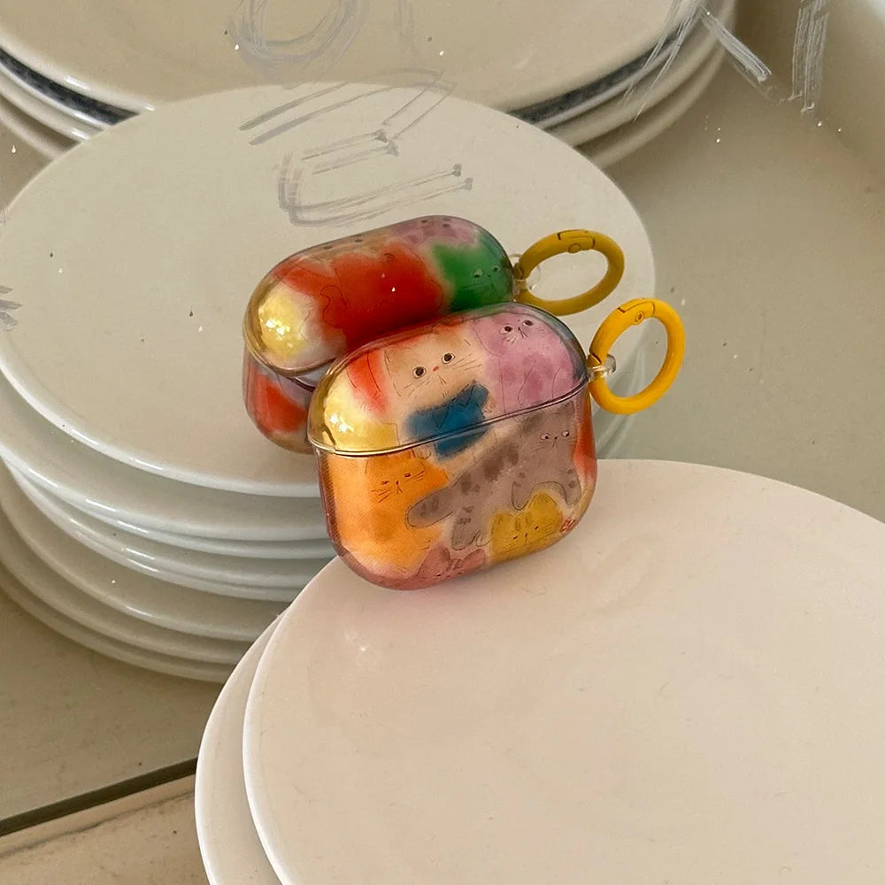 Two colorful, translucent AirPods cases with yellow keychain rings, featuring a marbled tie-dye pattern in orange, pink, blue, green, and yellow, resting on stacked white ceramic plates.