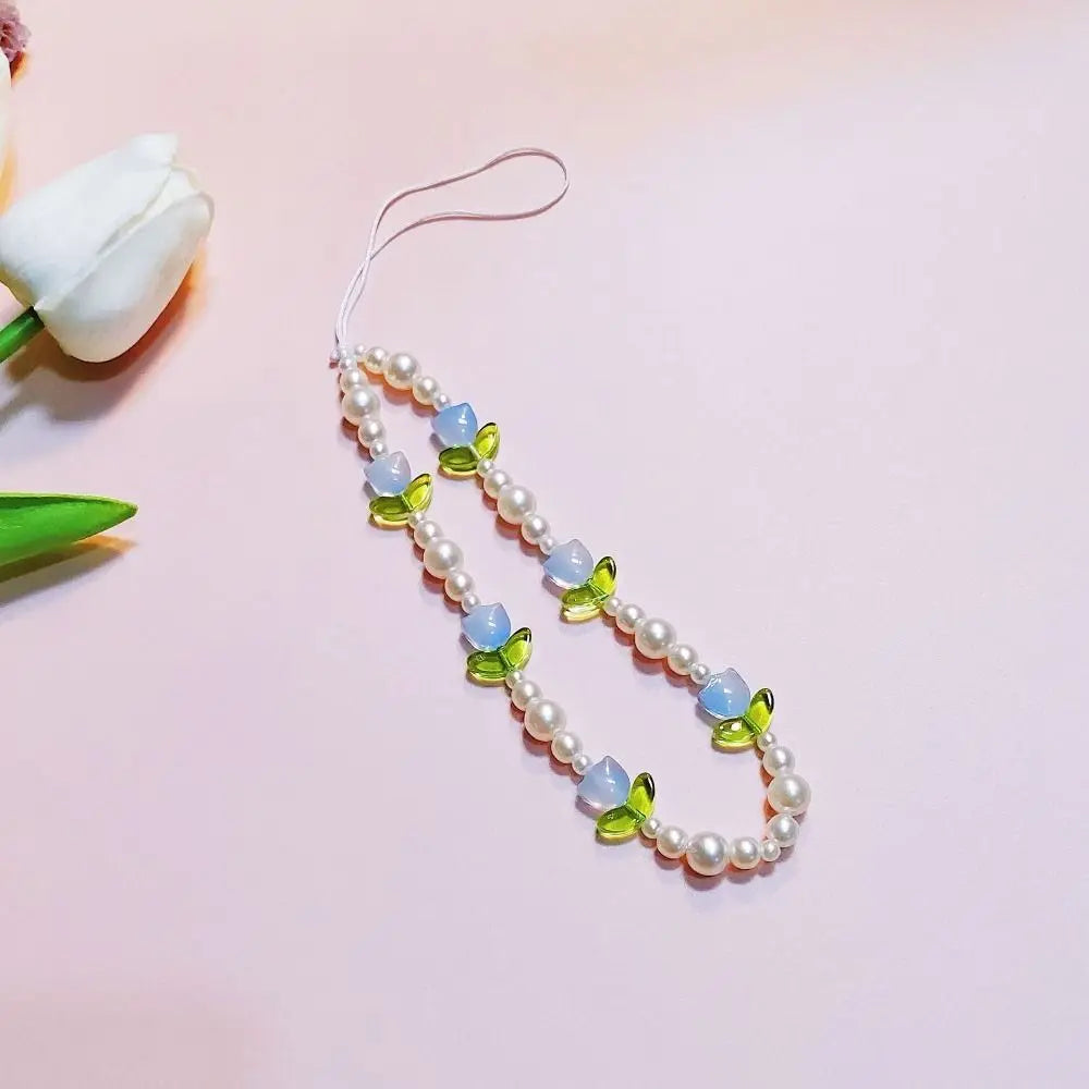 A beaded phone strap or necklace featuring white faux pearls, light blue flower-shaped beads, and green leaf-shaped beads, displayed on a soft pink background with a white tulip in the corner.