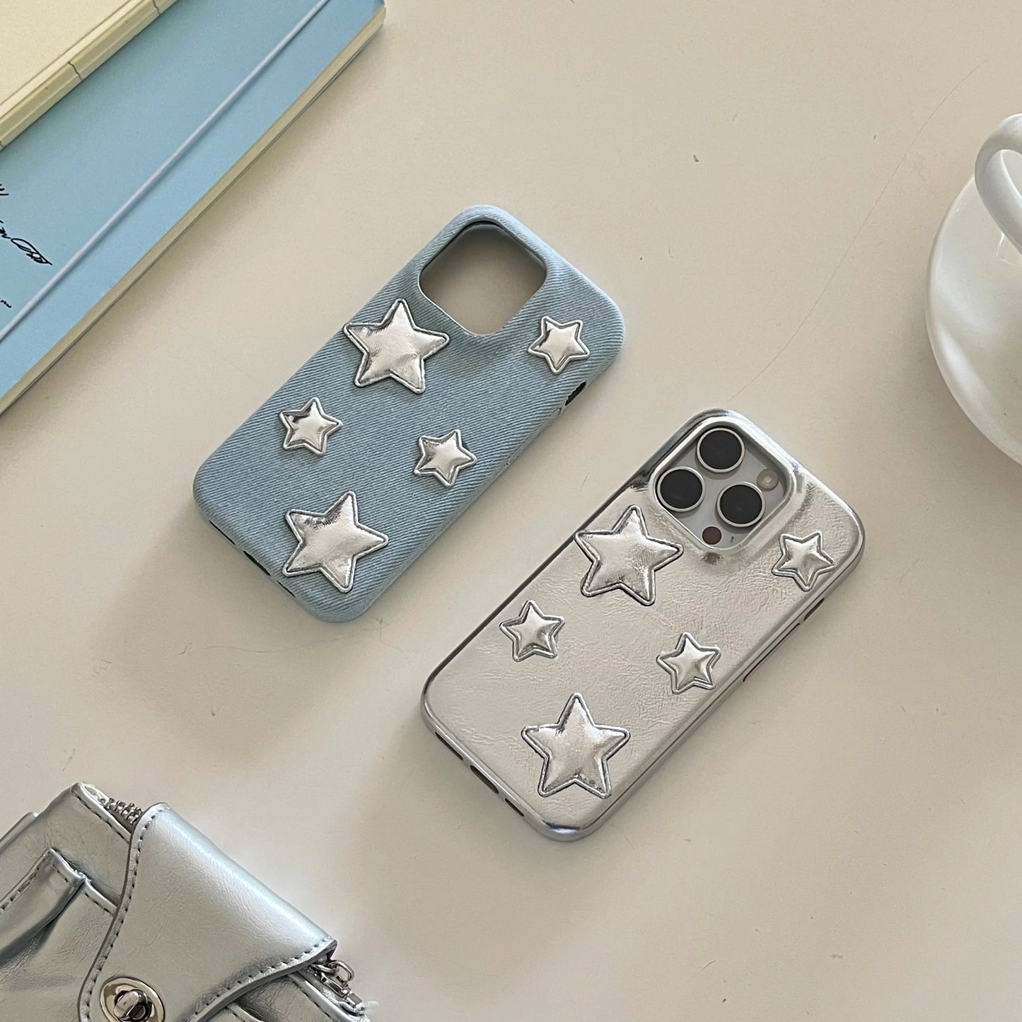 Two phone cases with raised star embellishments, one light blue denim-textured and one metallic silver, displayed on a beige tabletop with partial props including a book, cup, and small metallic pouch.