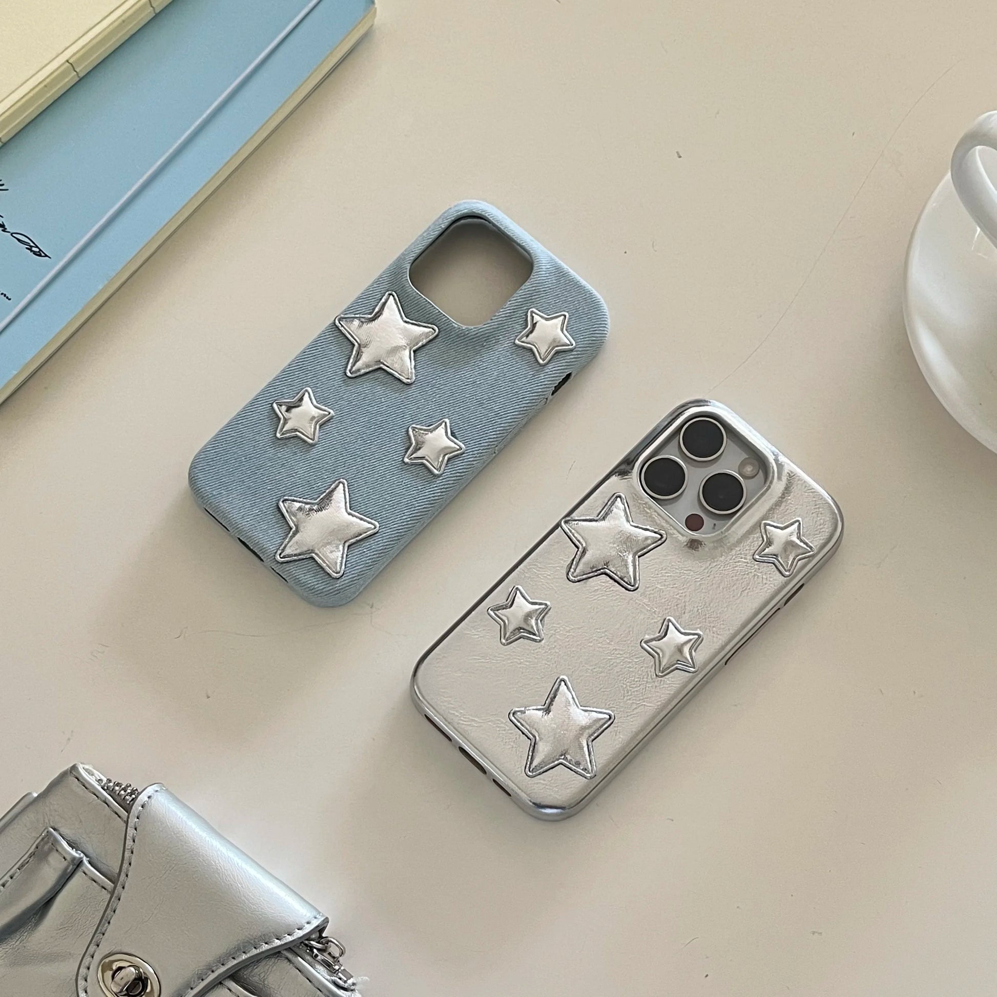 Two phone cases with raised star embellishments, one light blue denim-textured and one metallic silver, displayed on a beige tabletop with partial props including a book, cup, and small metallic pouch.