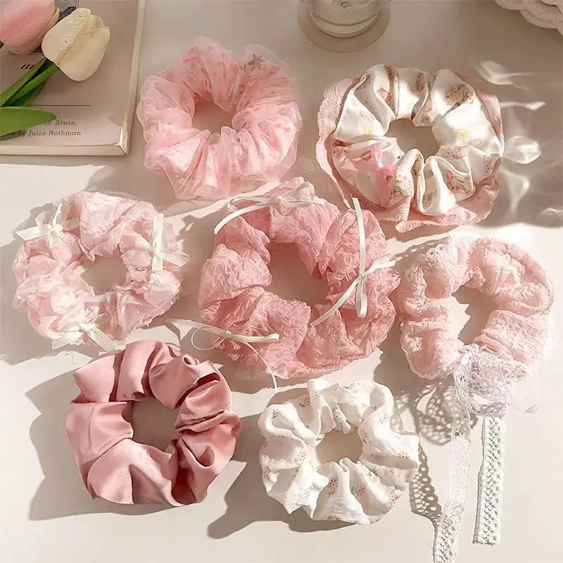 Six pink and white hair scrunchies arranged on a light beige surface with soft shadows. The set includes sheer pink mesh, textured pink lace, satin pink, and floral white scrunchies with ribbon ties and lace trim.