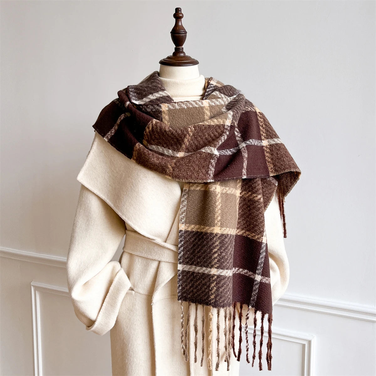A brown and beige plaid scarf with fringe tassels, draped over a cream-colored coat on a mannequin against a light gray wall.